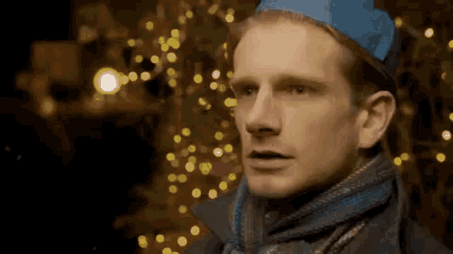 a man wearing a blue hat and scarf is standing in front of a christmas tree with lights .