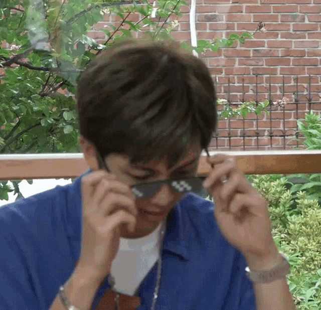 a man in a blue shirt is adjusting his sunglasses with a brick wall in the background .