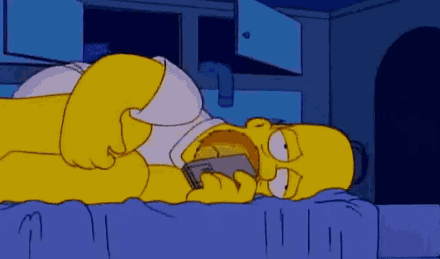 homer simpson is laying on a bed with his mouth open while holding a cell phone .