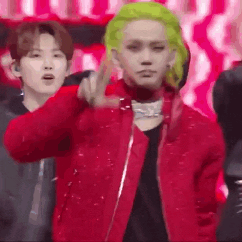 a man with green hair is wearing a red jacket and making a peace sign .