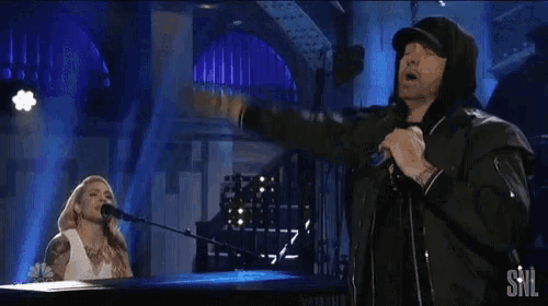 a man is singing into a microphone while a woman plays a piano on stage .