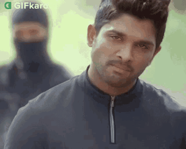 a man in a black jacket is looking at the camera while a man in a black mask stands behind him .