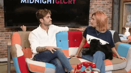 a man and a woman sit on a colorful couch in front of a midnight glory tv