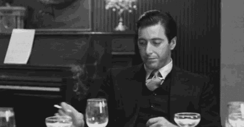 a man in a suit and tie is sitting at a table with wine glasses and a cigarette in his hand