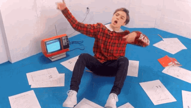 a man in a plaid shirt is sitting on the floor surrounded by papers and a tv