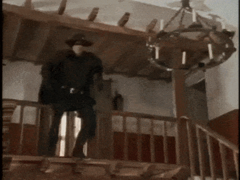 a man in a mask is standing on a balcony in a room with a chandelier .