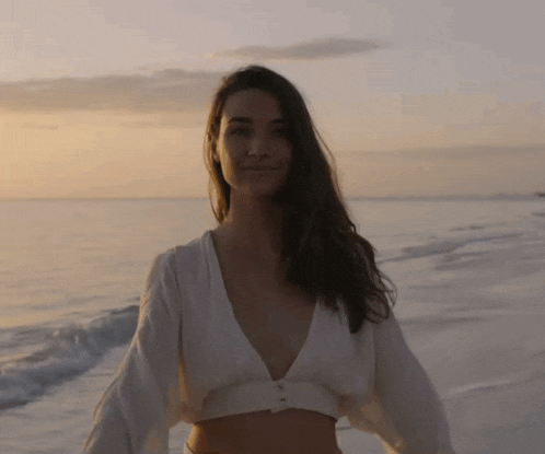 a woman in a white top is walking on the beach at sunset