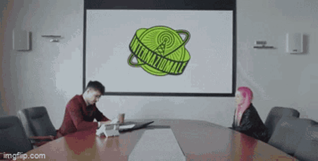 a man and a woman sit at a conference table in front of a large screen that says international