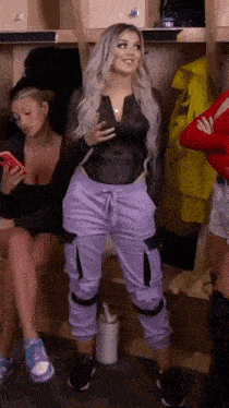 a woman in purple pants and a black top is standing in a locker room with other women .