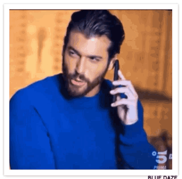 a man with a beard is talking on a cell phone while wearing a blue sweater .