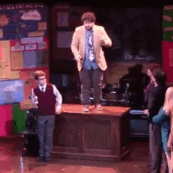 a man is standing on top of a wooden desk on a stage in front of a group of people .
