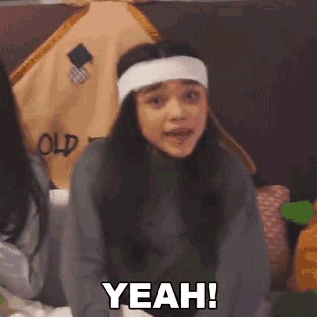 a woman wearing a headband and a gray sweater is making a funny face and saying yeah .