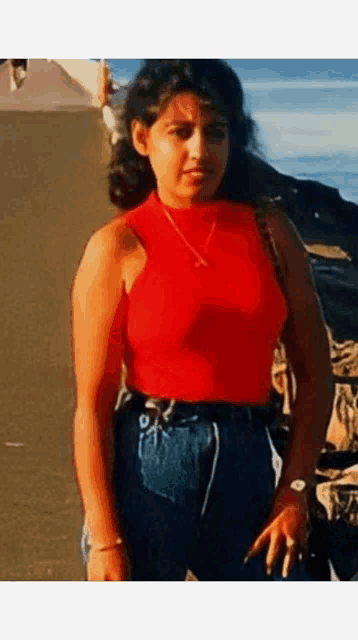 a woman in a red tank top and blue jeans stands on a beach