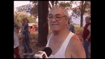 a man in a white tank top is talking into a microphone while standing in front of a group of people .
