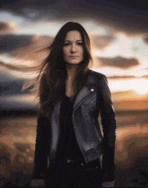 a woman in a black leather jacket stands in a field at sunset