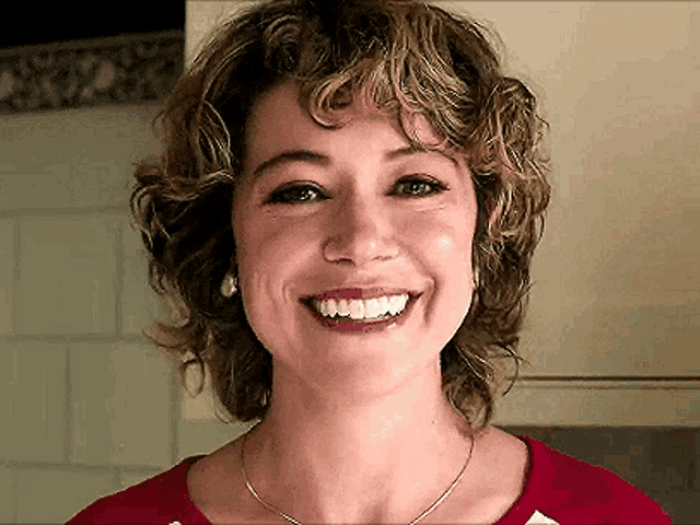 a woman with curly hair is smiling and wearing a red shirt and necklace