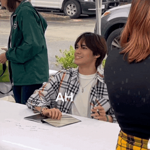 a man in a plaid shirt sits at a table with ah written on his shirt