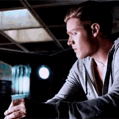 a man in a gray hoodie is sitting at a table in a dark room looking at something .