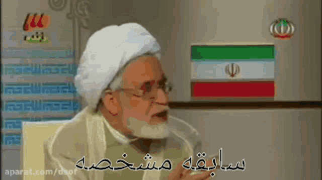 a man with a beard and glasses is talking in front of a refrigerator with a foreign flag on it