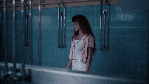a woman in a white tank top stands in a bathroom with blue tiles