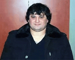 a man in a black coat is standing in front of a wooden door and looking at the camera .