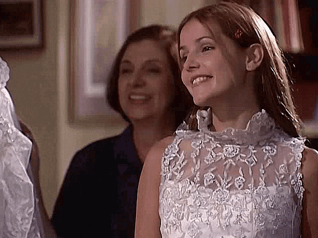 a young girl is wearing a white wedding dress and smiling while standing next to a woman .