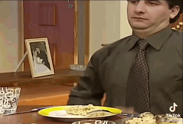 a man in a tie is sitting at a table with a plate of food and a picture of a bride and groom .