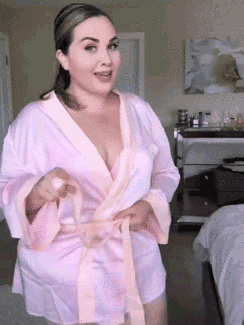a woman in a pink robe is standing in a bedroom and pointing at the camera .