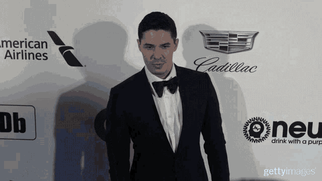 a man in a tuxedo is standing in front of a wall with logos for american airlines cadillac and oneu