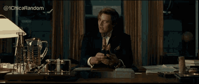 a man in a suit and tie is sitting at a desk and looking at his phone