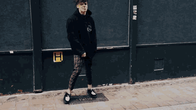 a man wearing a black hoodie and plaid pants stands on the sidewalk