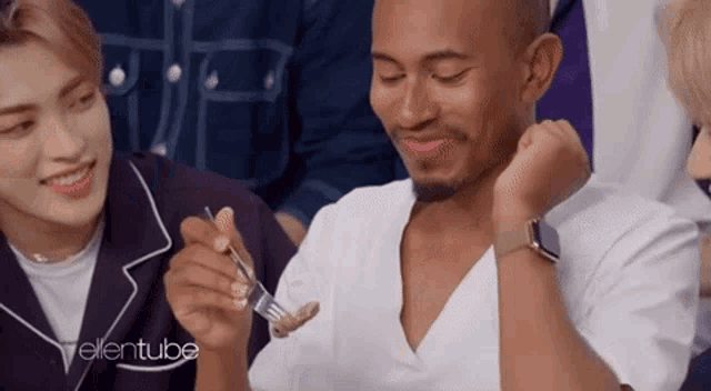 a man is eating food with a fork and smiling while another man watches .