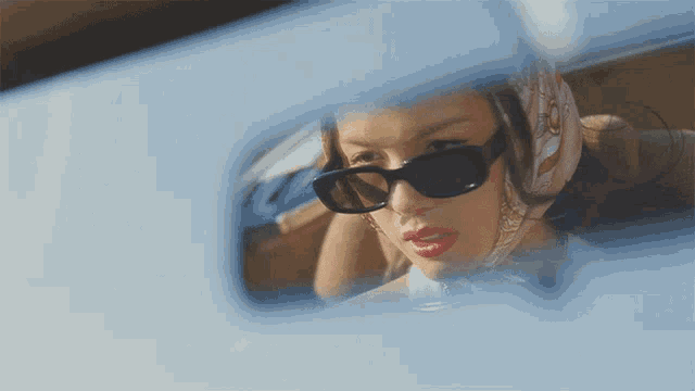 a woman wearing sunglasses and a scarf looks at herself in the rear view mirror of a car