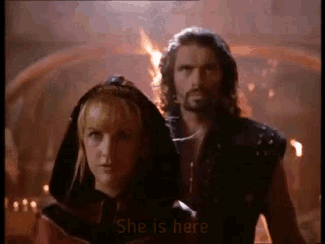 a man and a woman are standing next to each other in front of a fire and the woman is saying `` she is here '' .