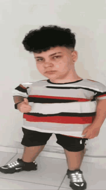 a young boy wearing a red white and black striped shirt and black shorts giving a thumbs up