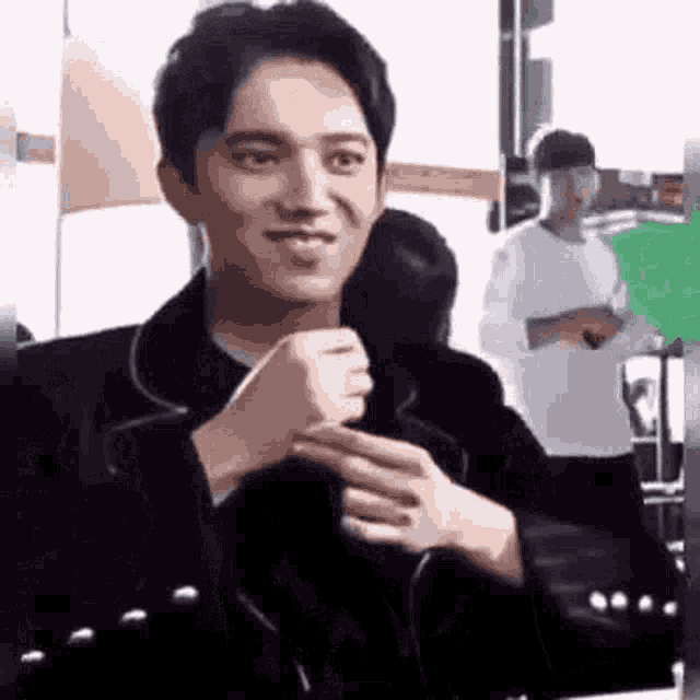 a man in a black jacket is sitting in a chair with his hands folded and smiling .