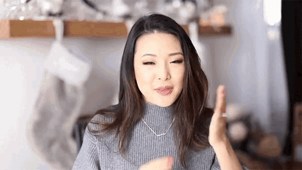 a woman wearing a gray sweater and a necklace is making a funny face and waving her hand .