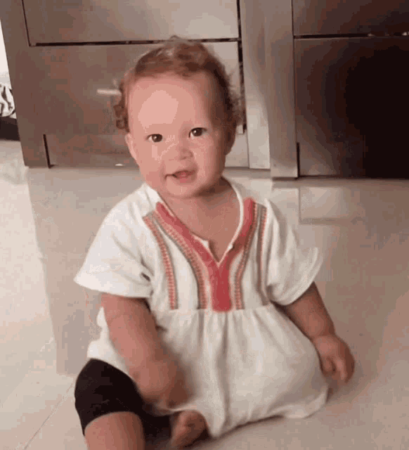 a baby is sitting on the floor wearing a white dress with a red y on it