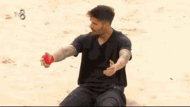 a man with a tattoo on his arm is sitting on the sand with his arms outstretched and a tv8 logo in the background .