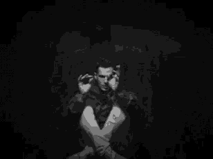 a black and white photo of a man sitting in a dark room with his hands on his face .