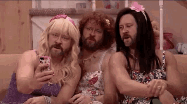 three bearded men are sitting on a couch taking a selfie with their cell phone .