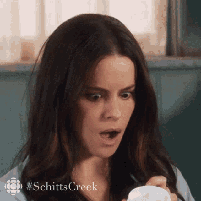 a woman with a surprised look on her face is holding a cup with #schitts creek written on it