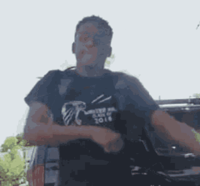 a man wearing a black shirt with the year 2016 on it is standing in front of a black suv .