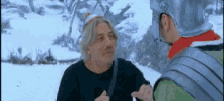 two men are standing in the snow talking to each other . one of the men is wearing a helmet .