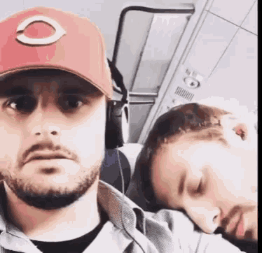 a man wearing a red hat and headphones is taking a selfie with another man sleeping on a plane .