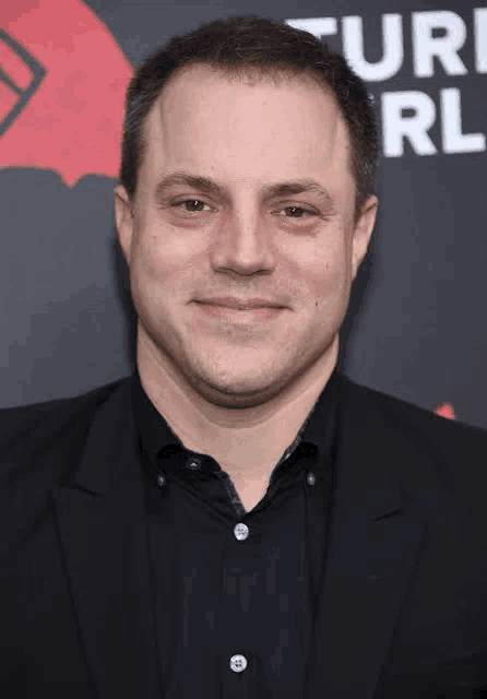 a man in a suit and black shirt is standing on a red carpet and smiling .