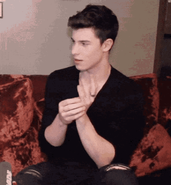 a young man in a black shirt is sitting on a couch with his hands together .