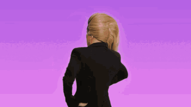 a drag queen wearing a black jacket and gold buttons stands on a purple background