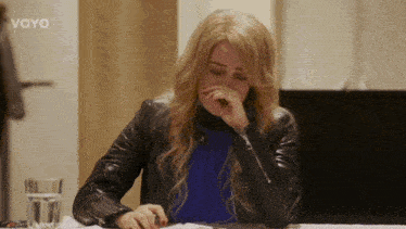 a woman in a leather jacket sits at a table with a glass of water and the word voyo on the bottom right