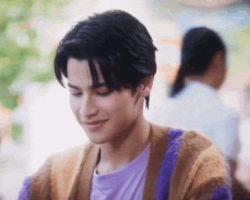 a young man wearing a purple t-shirt and a brown and purple cardigan is smiling .